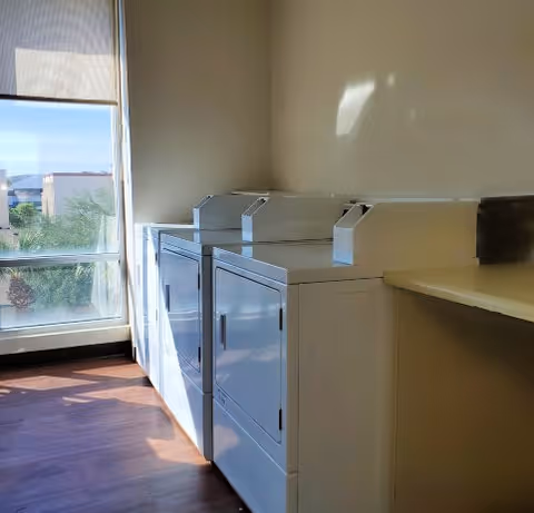 A laundry room with two white coin-operated washing machines or dryers placed side by side against a beige wall. There is a large window on the left side letting in natural light, and a countertop on the right side of the machines. The floor is made of wood or wood-like material.
