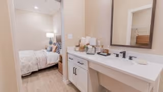View of a bedroom and bathroom vanity area in a senior living facility. The bedroom has a neatly made bed with pillows and a bedside lamp. The bathroom area features a white countertop with a sink, black faucet, mirror, and various toiletries arranged on the counter.