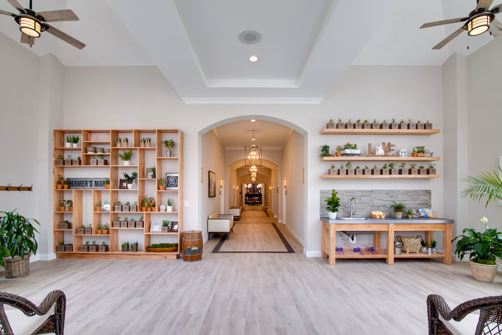 Bright and spacious interior hallway with light wood flooring and white walls. On the left, a large wooden shelving unit filled with small potted plants and decorative items. On the right, a wooden counter with a sink, more potted plants, and shelves above holding small potted plants and decorative letters spelling 'LOVE'. Ceiling fans with lights are visible, and the hallway extends into the distance with multiple light fixtures and seating along the walls.