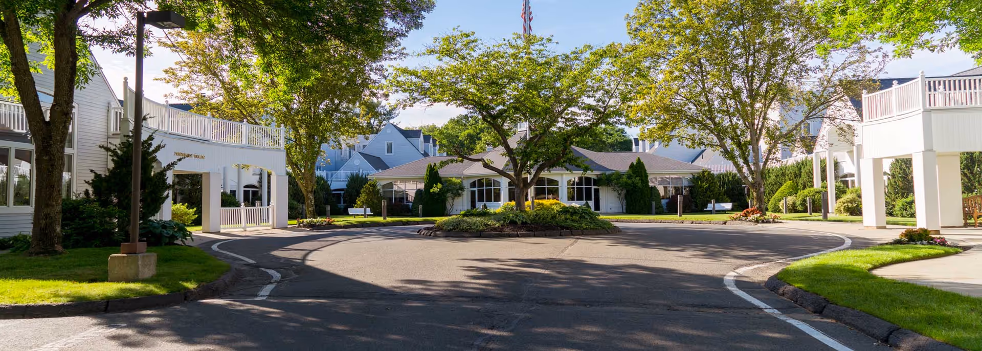 Entrance area of Pomperaug Woods senior living facility with a circular driveway, landscaped greenery, trees, and white buildings under a clear blue sky.