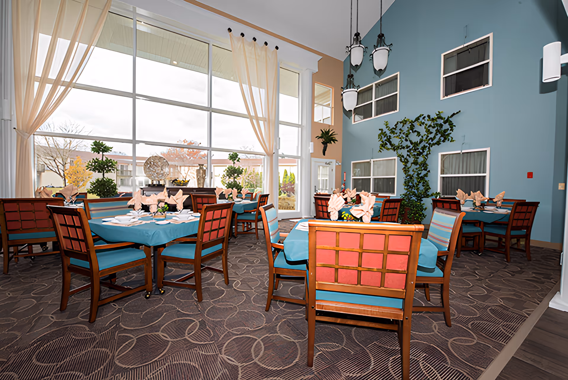 A bright dining room with large floor-to-ceiling windows letting in natural light. The room features multiple tables covered with teal tablecloths, each set with folded napkins and tableware. The chairs have wooden frames with teal and red upholstery. The walls are painted in blue and beige tones, decorated with plants and hanging light fixtures.