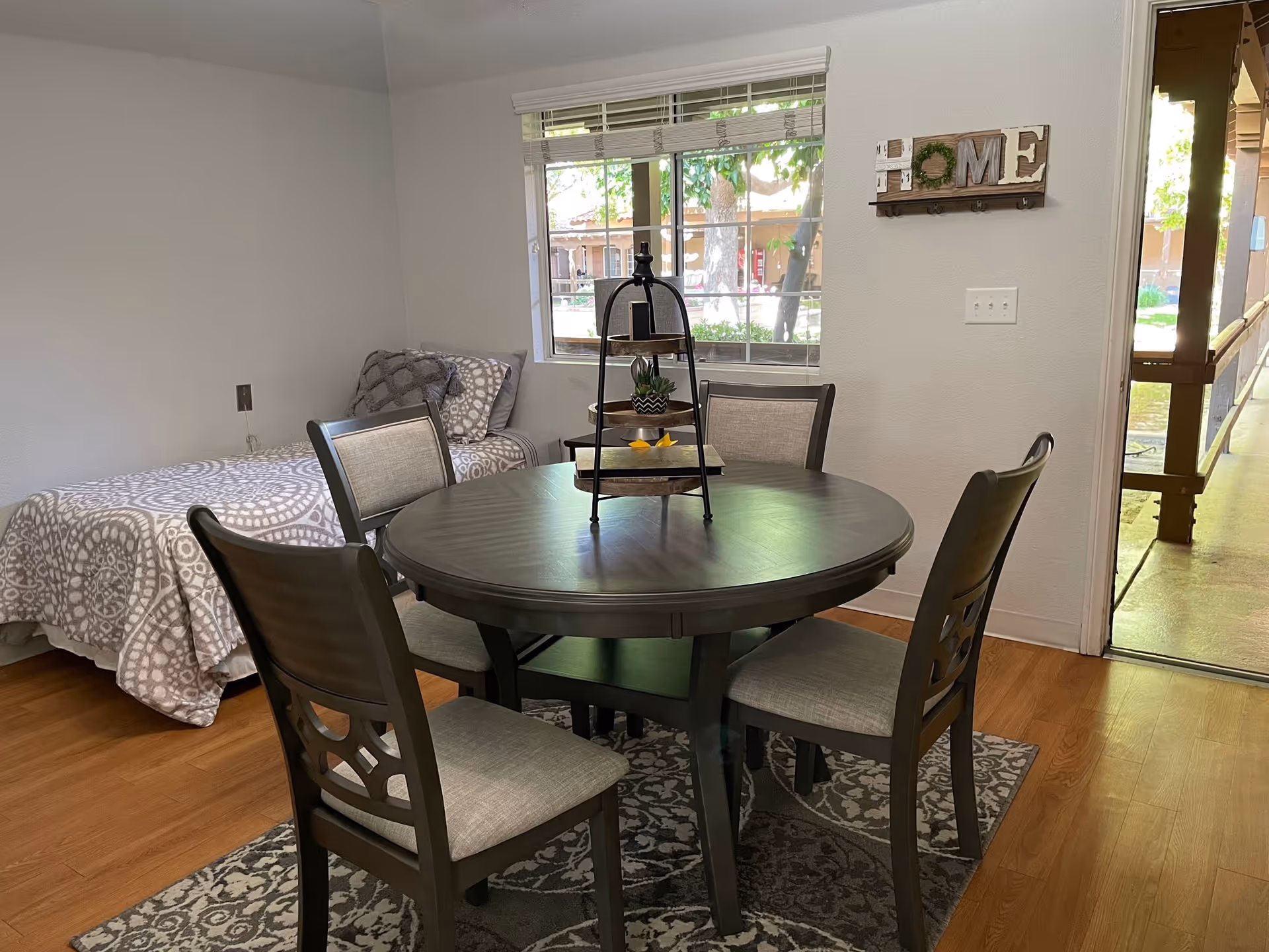 A cozy room with a round wooden dining table surrounded by four cushioned chairs on a patterned rug. In the background, there is a single bed with patterned bedding and pillows near a window with blinds. A decorative 'HOME' sign is mounted on the wall, and an open door leads to an outdoor walkway with wooden beams and greenery visible outside.