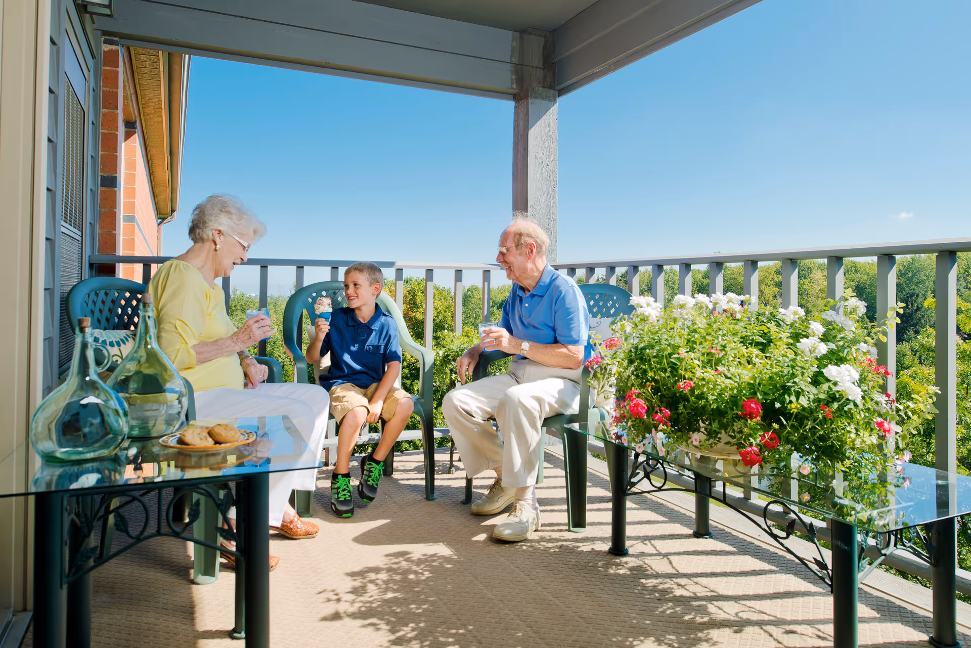 An elderly woman, a young boy, and an elderly man sitting on a balcony with green plastic chairs, enjoying ice cream and drinks. The balcony has a glass table with cookies and decorative glass bottles, and a large planter with blooming red and white flowers. The background shows a clear blue sky and green trees.