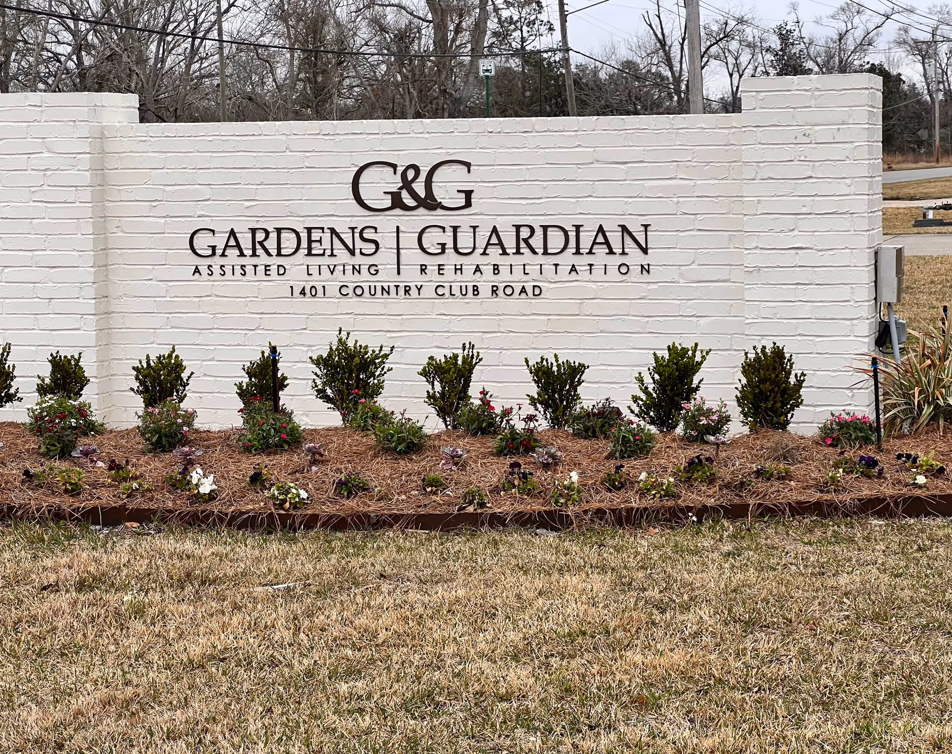 White brick entrance sign reading 'G&G Gardens | Guardian Assisted Living Rehabilitation' with planted shrubs and flowers in front.