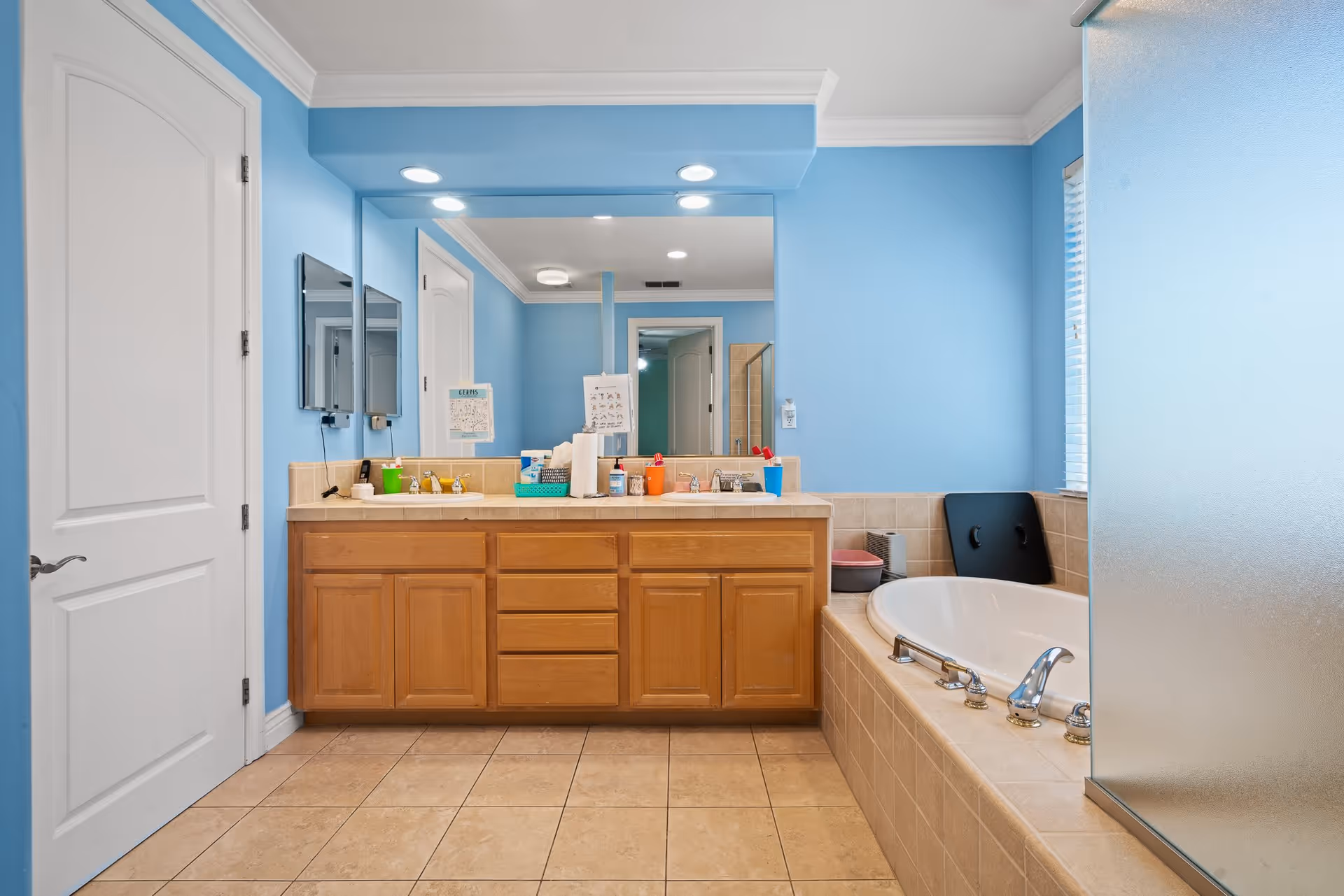 A bathroom with light blue walls, a double sink vanity with wooden cabinets, a large mirror above the sinks, and a tiled bathtub with chrome fixtures. There is a frosted glass shower door on the right side and a white door on the left. Various toiletries and cleaning supplies are placed on the vanity counter.