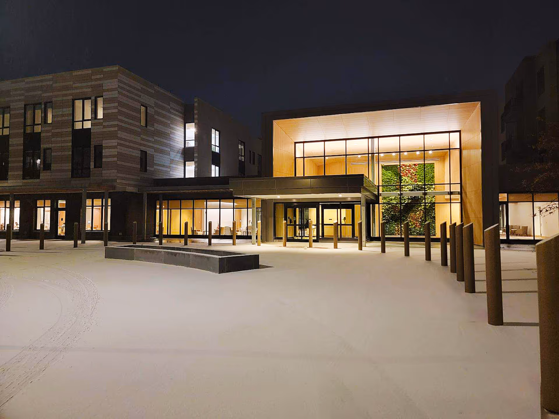 Night view of the exterior entrance of The Baldwin Senior Living facility, showing a modern building with large glass windows, illuminated interior, and a snow-covered ground in front.