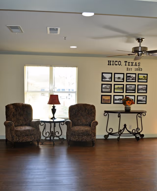 A sitting area with two patterned armchairs separated by a small round table with a red lamp on it. Behind the chairs is a window with blinds. On the right side, there is a decorative table with a flower arrangement and a wall display of framed black and white photos with the text 'HICO, TEXAS Est 1883' above them. The floor is wooden, and a ceiling fan is visible above.