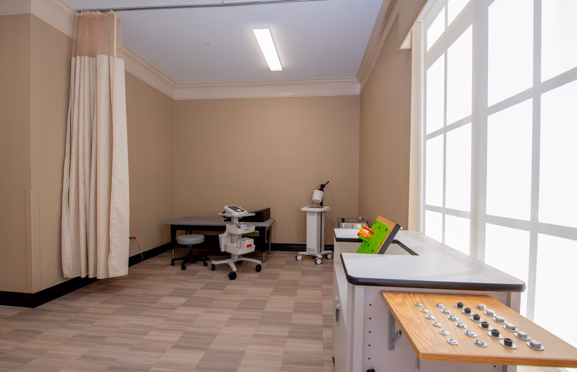 A medical examination or therapy room with beige walls and wood-patterned flooring. The room contains a medical examination table with a stool, a mobile medical cart with equipment, a fire extinguisher on a small stand, and a therapy device with knobs and buttons on a counter near a large window with frosted glass panels. A beige privacy curtain is partially drawn on the left side.