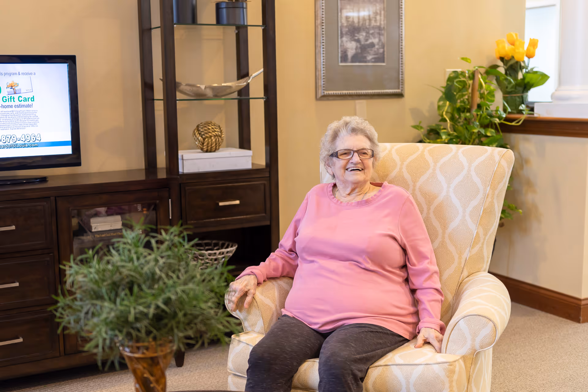 An elderly woman wearing glasses and a pink long-sleeve shirt sits smiling in a patterned beige armchair in a cozy living room. Behind her is a wooden cabinet with a TV on top, decorative items on glass shelves, a framed picture on the wall, and a window sill with yellow flowers and green plants.