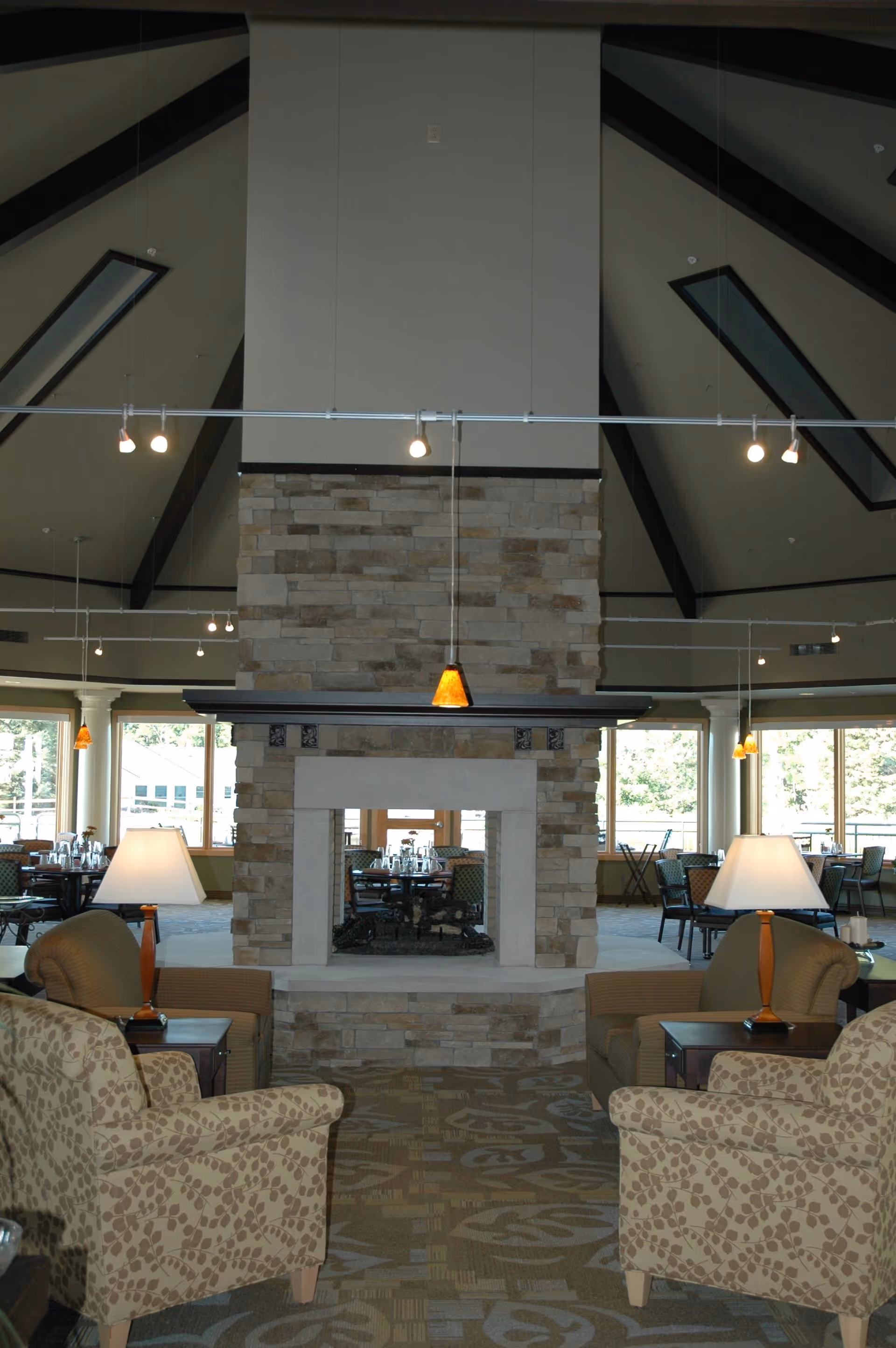 Interior view of a senior living facility lounge area featuring a large stone fireplace in the center. There are two patterned armchairs and two solid-colored armchairs arranged around the fireplace, each with a side table and lamp. The ceiling is vaulted with exposed dark beams and skylights. In the background, there are dining tables and chairs near large windows letting in natural light.