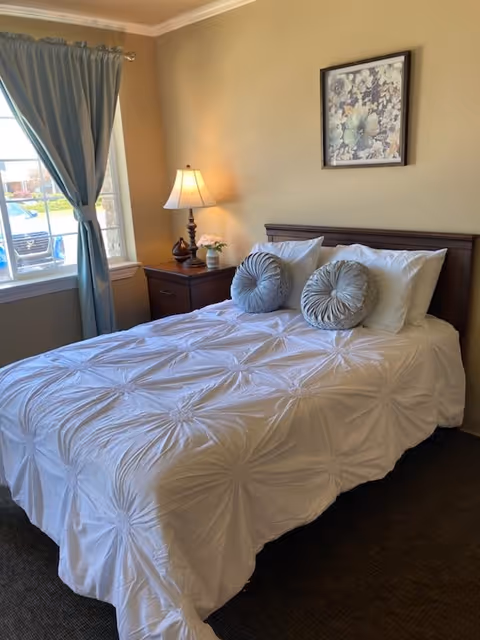 A neatly made bed with a white textured comforter and two round decorative pillows in a bedroom. Next to the bed is a wooden nightstand with a lamp and a small flower vase. A window with blue curtains is on the left side, and a framed floral artwork hangs on the beige wall above the bed.