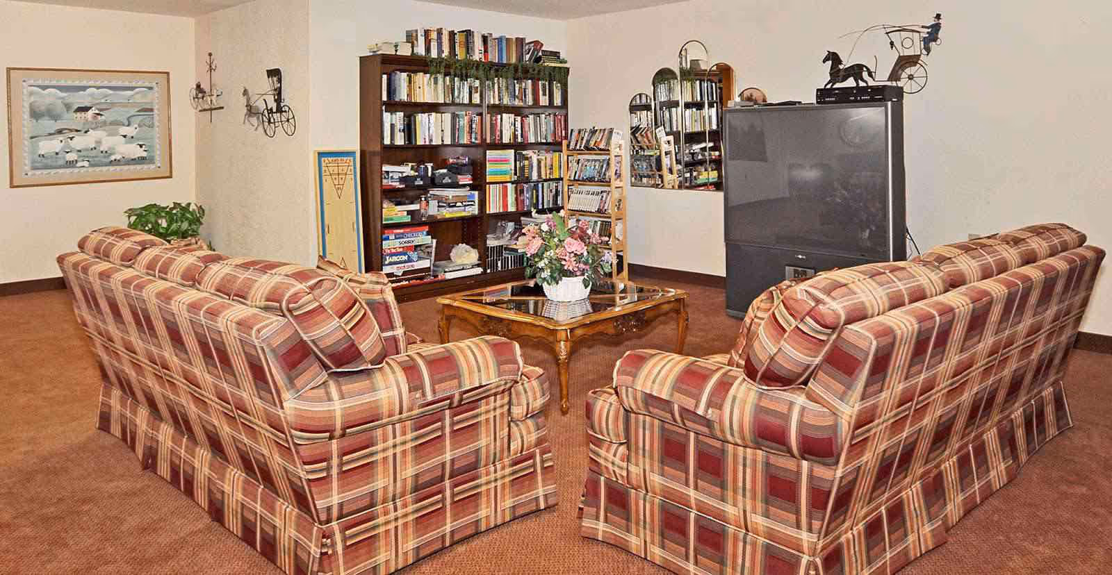 Two plaid sofas face a glass-top coffee table, bookshelf, and large TV in a cozy common living room.