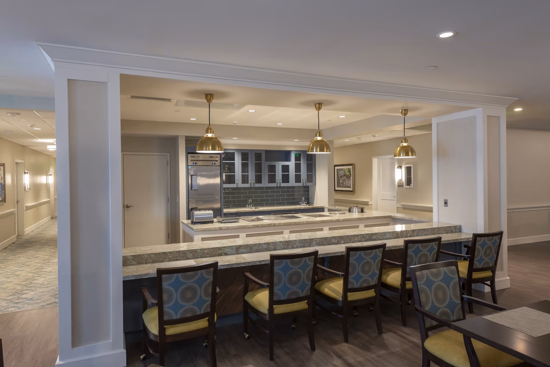 Interior view of a modern kitchen area in a senior living facility with a long marble countertop bar and six chairs with patterned upholstery. The kitchen features stainless steel appliances, glass-front cabinets, and three hanging brass pendant lights. There is a hallway visible to the left and framed artwork on the walls.