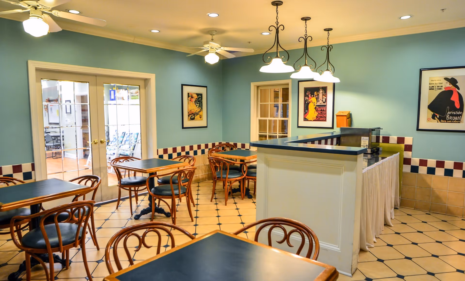 A cozy dining room with several wooden tables and chairs with dark blue tabletops and seats. The walls are painted light blue with a decorative tile border in red, white, and blue. There are three framed vintage-style posters on the walls. The room has ceiling fans with lights and a small counter area with a coffee machine. French doors lead to an adjacent room with additional seating.