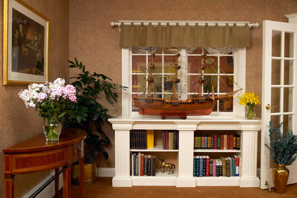 A cozy interior corner featuring a white built-in bookshelf filled with books and decorative items, including a model wooden ship on top. To the left, there is a wooden side table with a glass vase holding pink and white flowers, and a large green plant behind it. On the right, a glass door with white framing is partially open, and a golden vase with green foliage is placed on the floor. The walls are covered in a textured brown wallpaper, and a framed painting hangs above the side table.