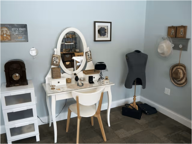 A cozy room corner featuring a white vanity table with an oval mirror, a white chair, and various decorative items including framed photos, a small lamp, and jewelry. To the left is a small white storage unit with an antique radio on top. On the right side, there is a gray dress form mannequin, a small shelf with two framed pictures, and two hats hanging on hooks on a light blue wall.