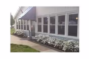 Exterior view of a single-story building with multiple windows, a covered entrance, and a flower bed with white flowers along the foundation. A sidewalk runs alongside the building.