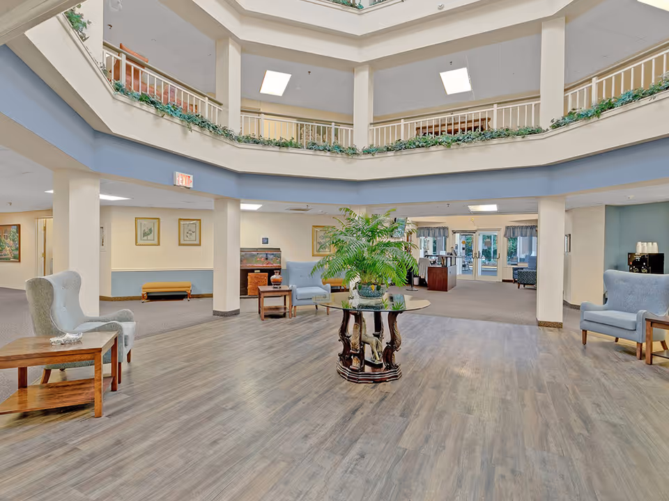 Spacious and well-lit senior living facility lobby with a round glass table holding a large green plant in the center. The area features comfortable armchairs, wooden side tables, and a second-floor balcony with railings and greenery. The flooring is a mix of wood and carpet, and there are framed pictures on the walls.