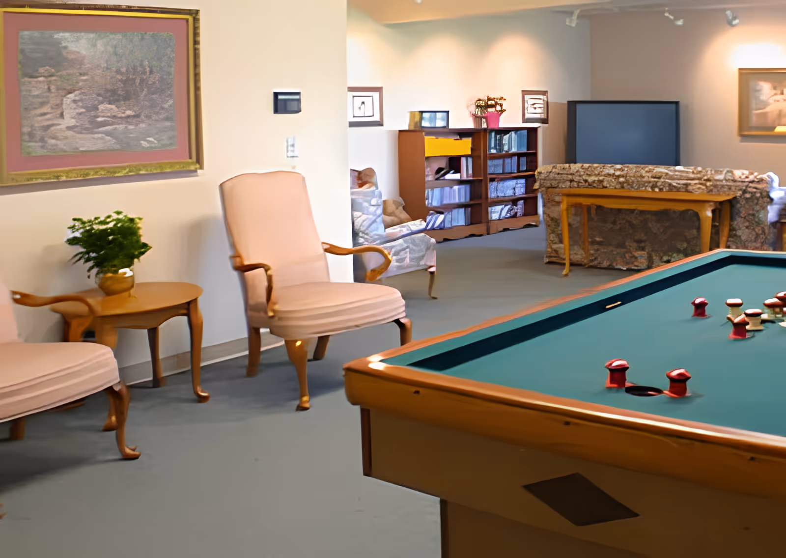 A cozy living room area with a pool table in the foreground, two upholstered armchairs with wooden legs and arms, a small wooden side table with a potted plant, a bookshelf filled with books and decorative items, and framed artwork on the walls.