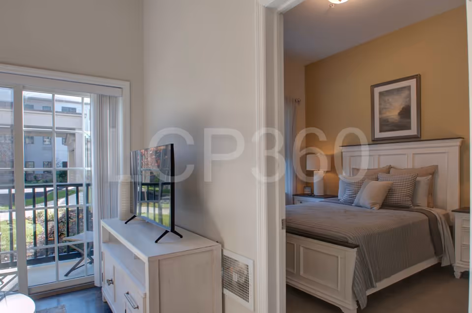 Interior view of a bedroom with a made bed and nightstand, adjacent to a TV on a console and a sliding glass door leading to a balcony.