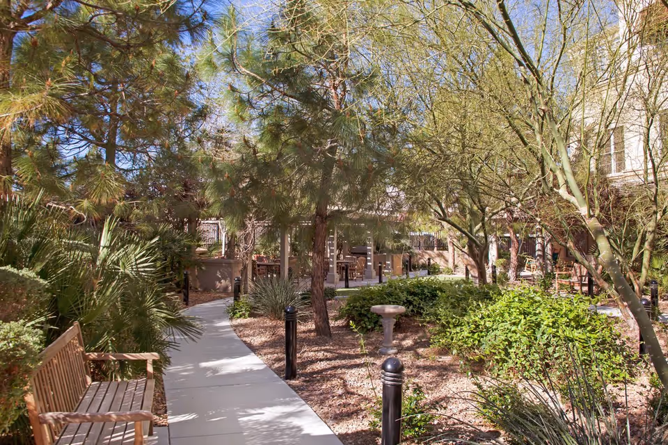 A sunny outdoor garden area at Sunrise of Henderson featuring a paved walkway, wooden benches, various green trees and shrubs, and a birdbath in the center. The garden is surrounded by a building and has seating areas under a pergola in the background.