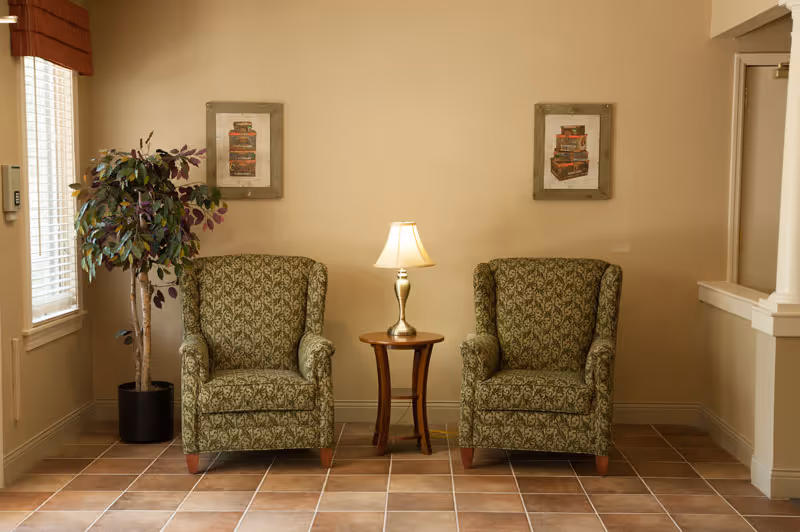 A cozy seating area with two patterned green armchairs separated by a small wooden side table with a lamp. There is a potted plant to the left near a window with blinds and two framed pictures on the beige wall behind the chairs. The floor is tiled with a brown and beige pattern.