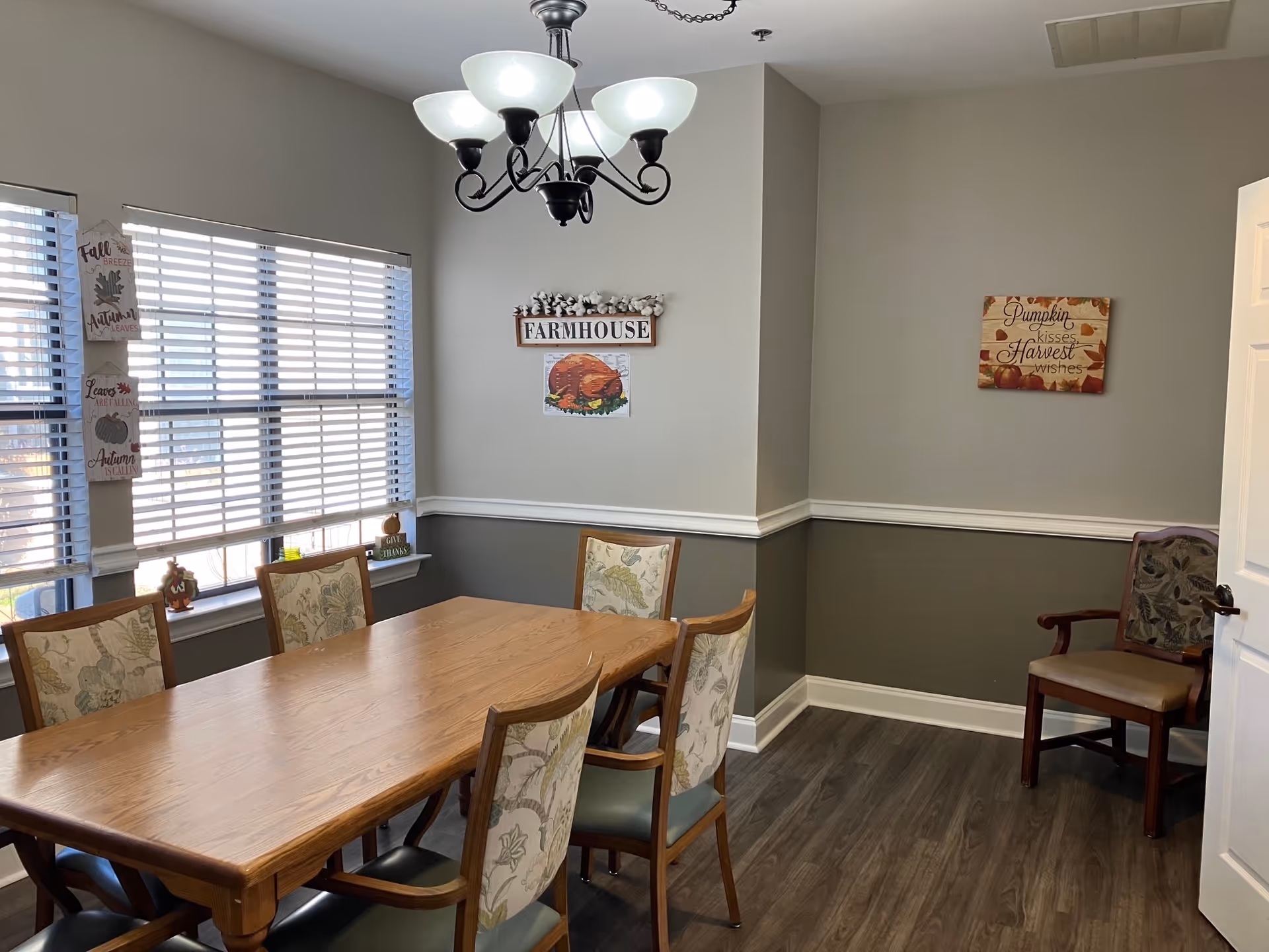 A dining room with a wooden table surrounded by six chairs with floral upholstery. The walls are painted in two tones, gray on the bottom and light beige on top, with white trim. There are three windows with white blinds on the left side, and fall-themed decorations on the walls including signs that say 'FARMHOUSE' and 'Pumpkin kisses Harvest wishes'. A chandelier with five frosted glass shades hangs above the table.