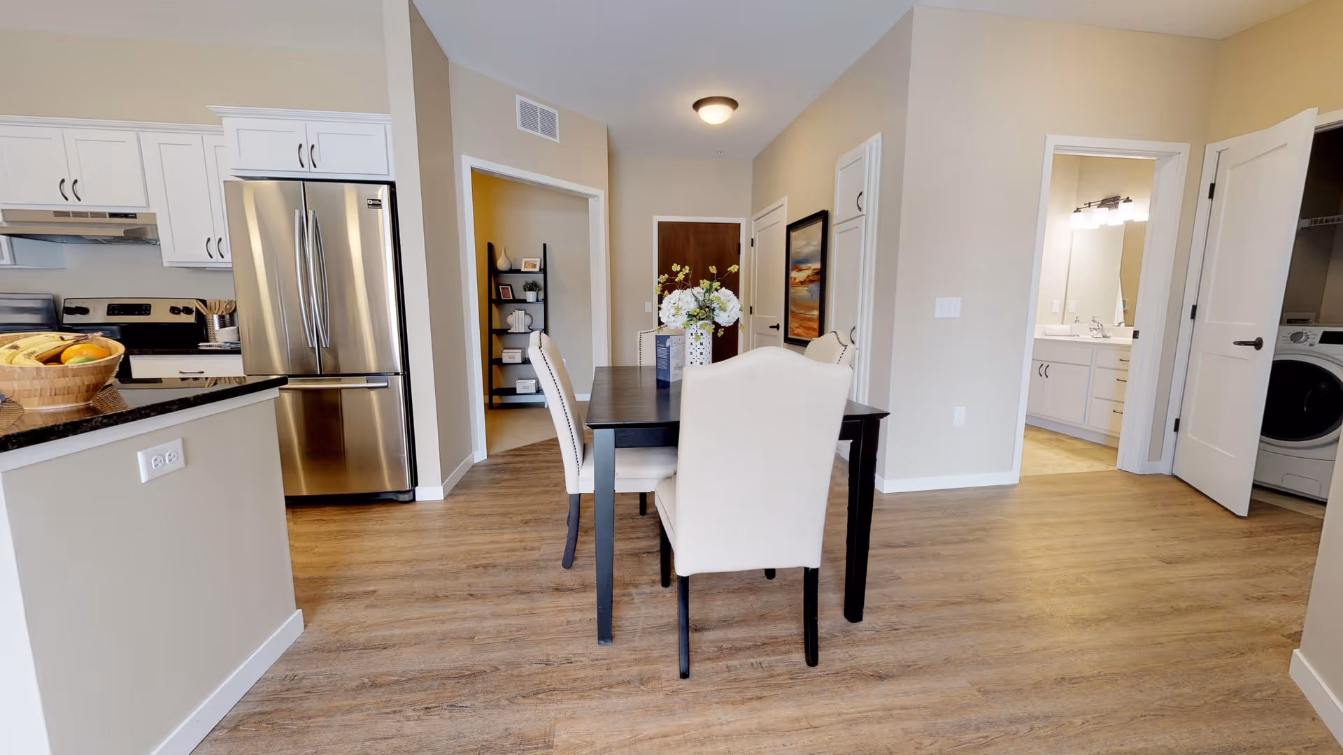 Interior view of a modern dining area with a dark wooden table and four beige upholstered chairs. To the left is a kitchen with white cabinets, a stainless steel refrigerator, and a countertop with a fruit basket. In the background, there is a hallway with a wooden door, a small shelving unit, and an open door revealing a bathroom with a sink and mirror. Another open door shows a laundry area with a washing machine.