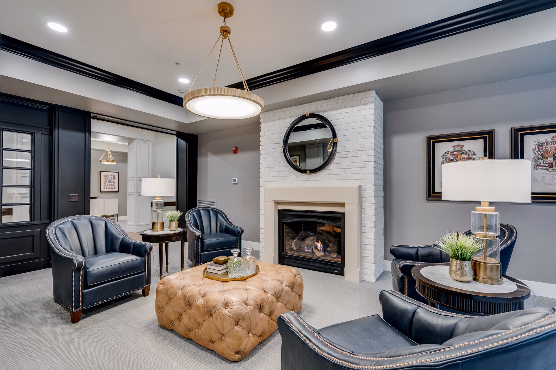 Cozy lounge with leather armchairs arranged around a tufted ottoman in front of a fireplace topped by a round mirror.