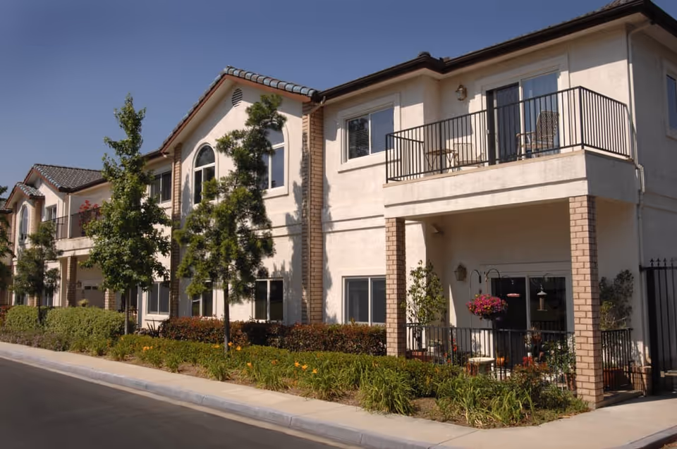 Exterior view of a two-story residential building with balconies and a patio, surrounded by small trees and landscaped greenery under a clear blue sky.