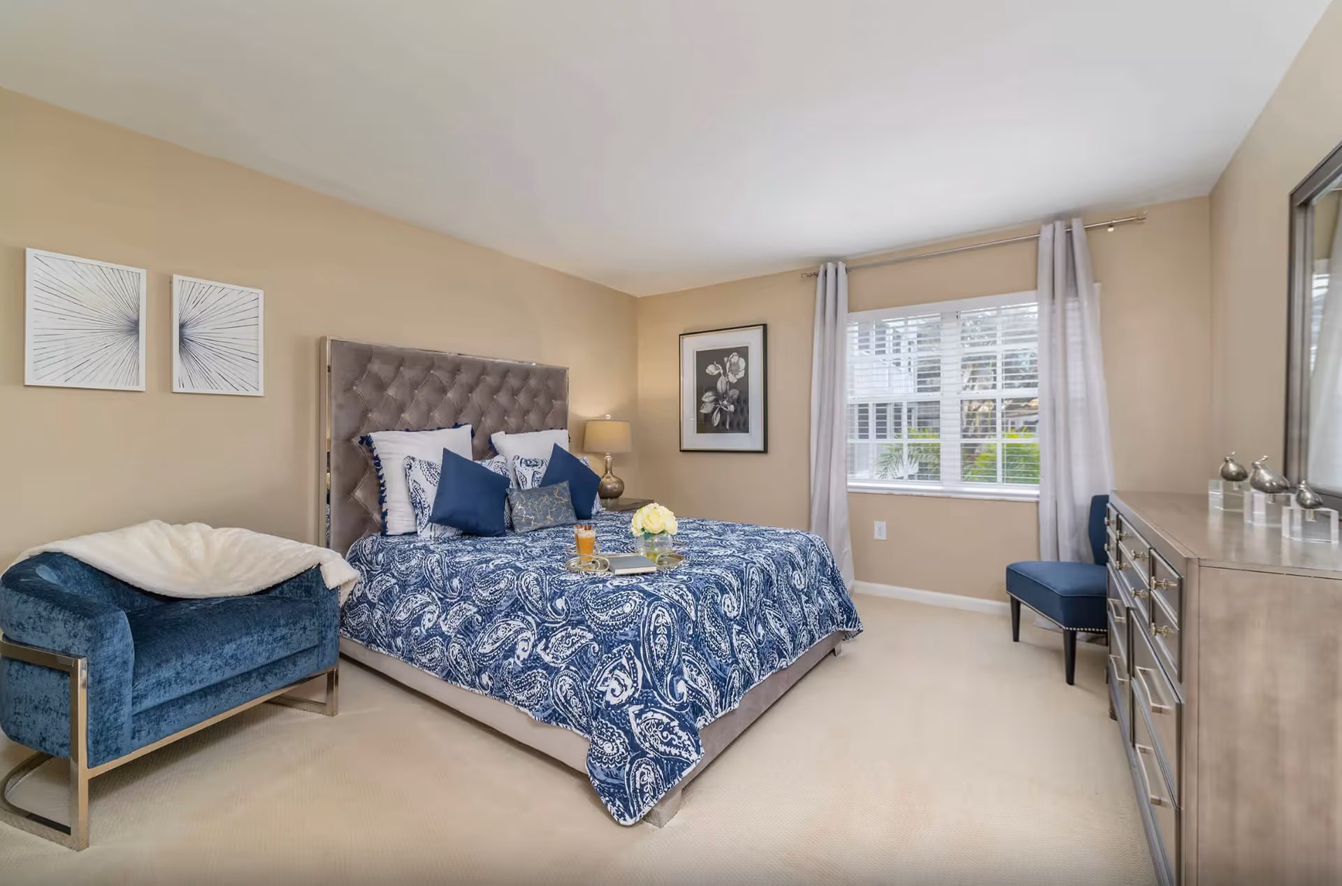 A well-lit bedroom with beige walls and carpet, featuring a large bed with a tufted gray headboard and blue paisley bedding. There are multiple pillows on the bed, a tray with a drink and flowers, a blue upholstered chair with a white throw blanket, a dresser with decorative items, a blue chair near the window with white curtains, and framed artwork on the walls.
