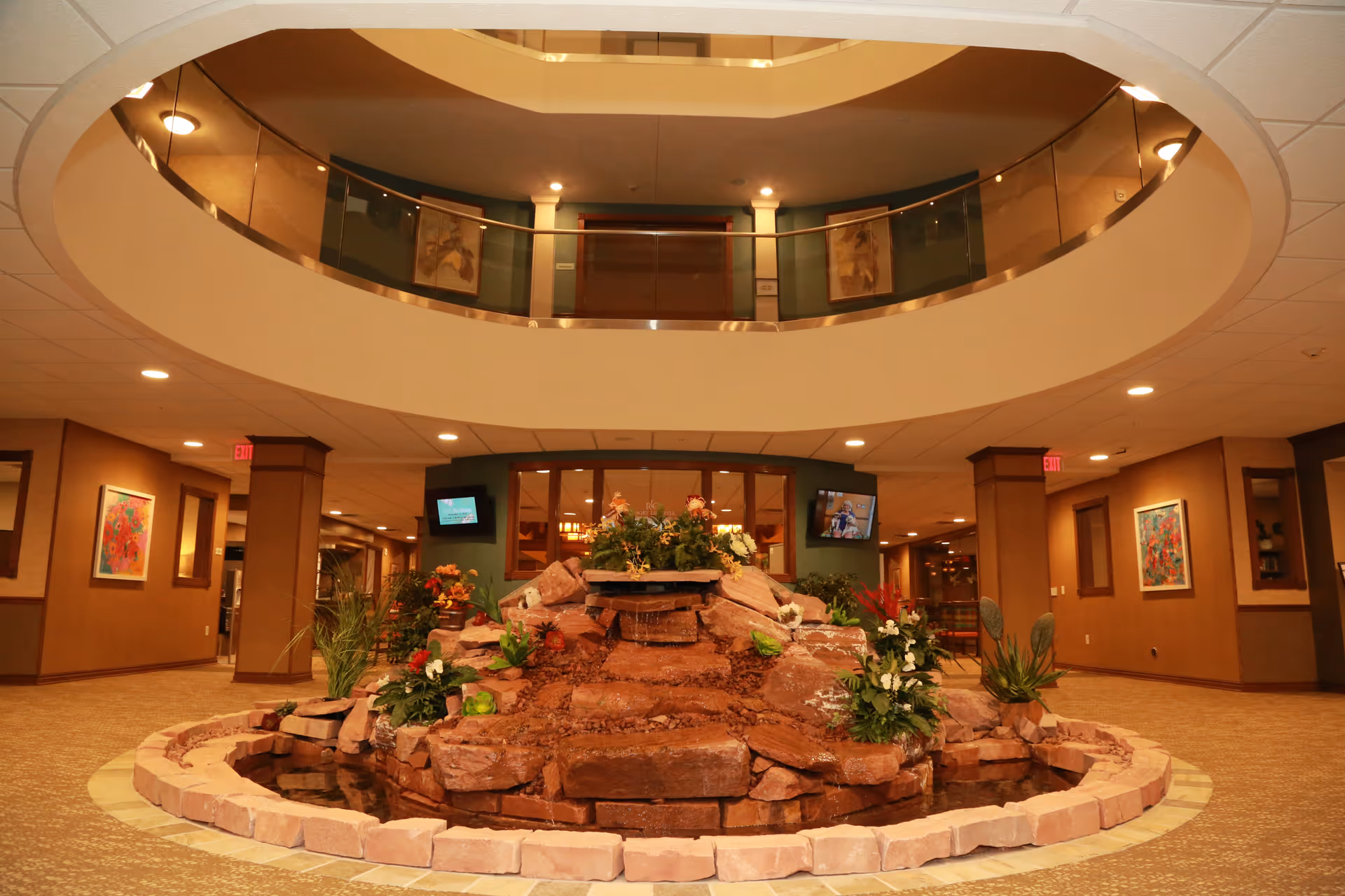 Interior view of a senior living facility lobby with a circular rock water feature decorated with plants and flowers in the center. The space has warm lighting, carpeted floors, and walls adorned with framed artwork. There is a second-floor balcony with glass railing overlooking the lobby area.