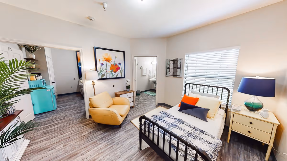 A bright and cozy assisted living room featuring a single bed with colorful pillows and a knitted blanket, a yellow armchair, a nightstand with a blue lamp, and a window with blinds. The room has wood flooring and light-colored walls. Visible through an open doorway is a small kitchen area with a turquoise refrigerator and microwave, and another doorway leads to a bathroom.