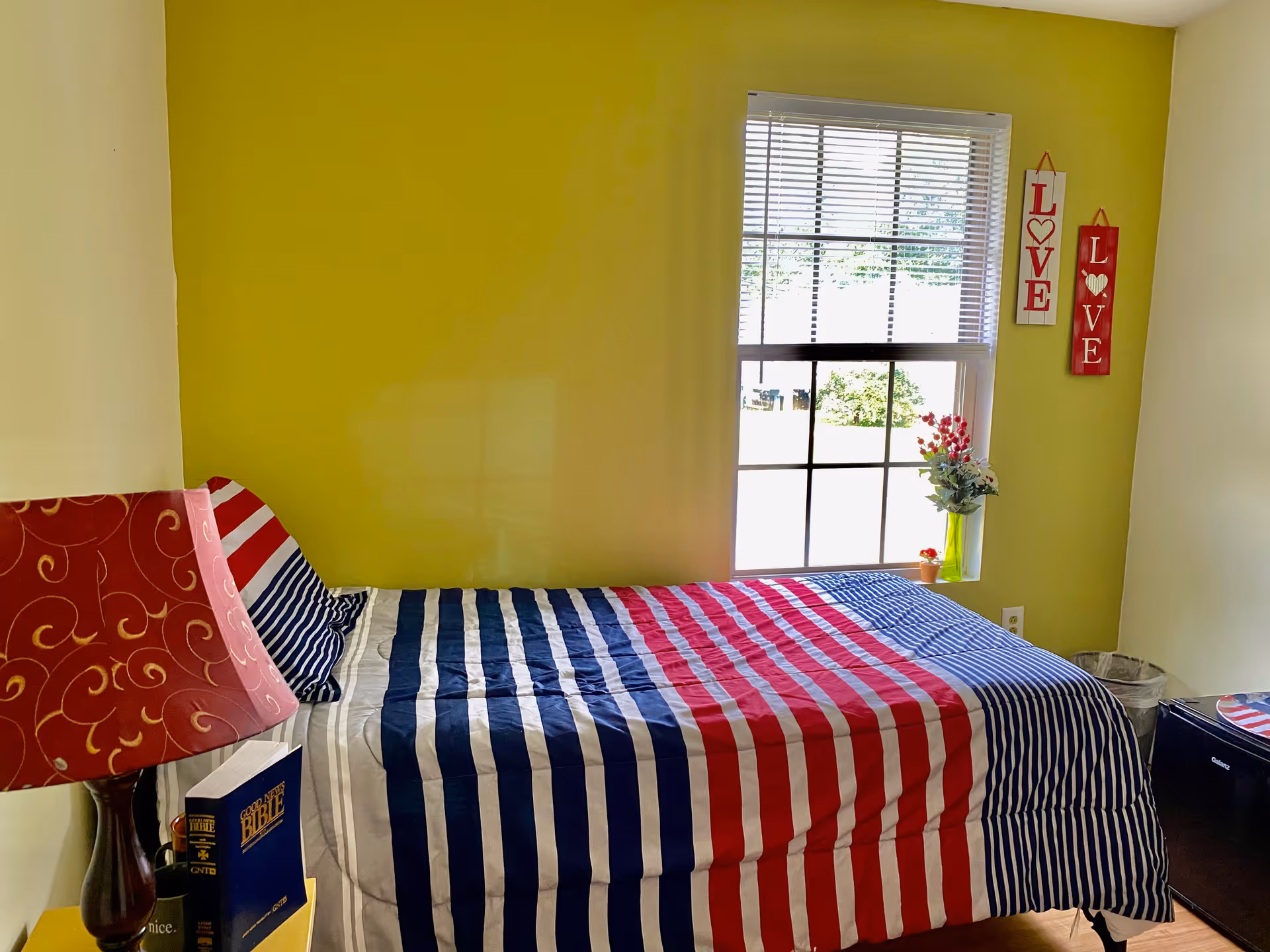 A bedroom with a bed covered in a striped red, white, and blue comforter. There is a window with blinds letting in natural light, two decorative signs on the wall that say 'LOVE', a small vase with flowers on the windowsill, a red lamp on a nightstand, and a blue book titled 'Good News Bible'.
