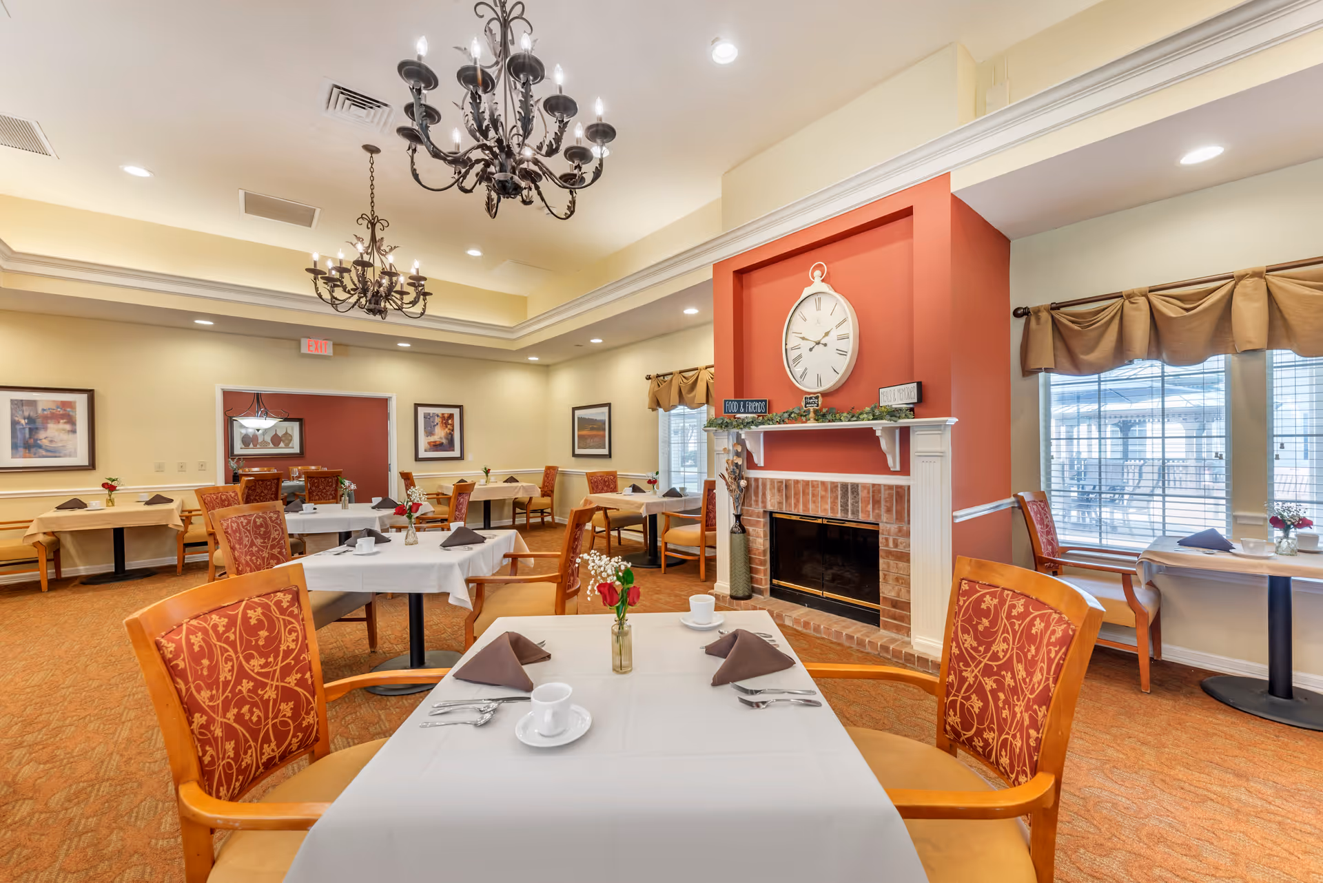A cozy dining room with several tables covered in white and beige tablecloths, each set with cups, napkins, and silverware. The room features red and gold patterned chairs, a brick fireplace with a large clock above it, and two ornate chandeliers hanging from the ceiling. Windows with brown valances allow natural light to enter the space.