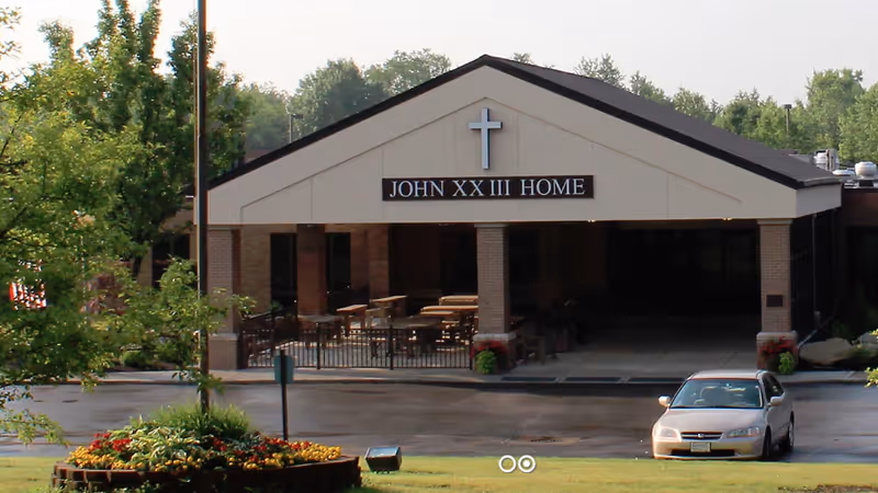 Exterior view of the entrance to Saint John XXIII Home, featuring a covered drop-off area with a cross and the name 'John XXIII Home' displayed above. There is a parked car in front and landscaping with trees and flowers around the driveway.