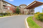 Exterior view of a senior living facility named The Summit, featuring a curved driveway leading to the main entrance with a covered drop-off area. The building has a combination of stone and red siding with multiple windows and landscaped greenery along the driveway.