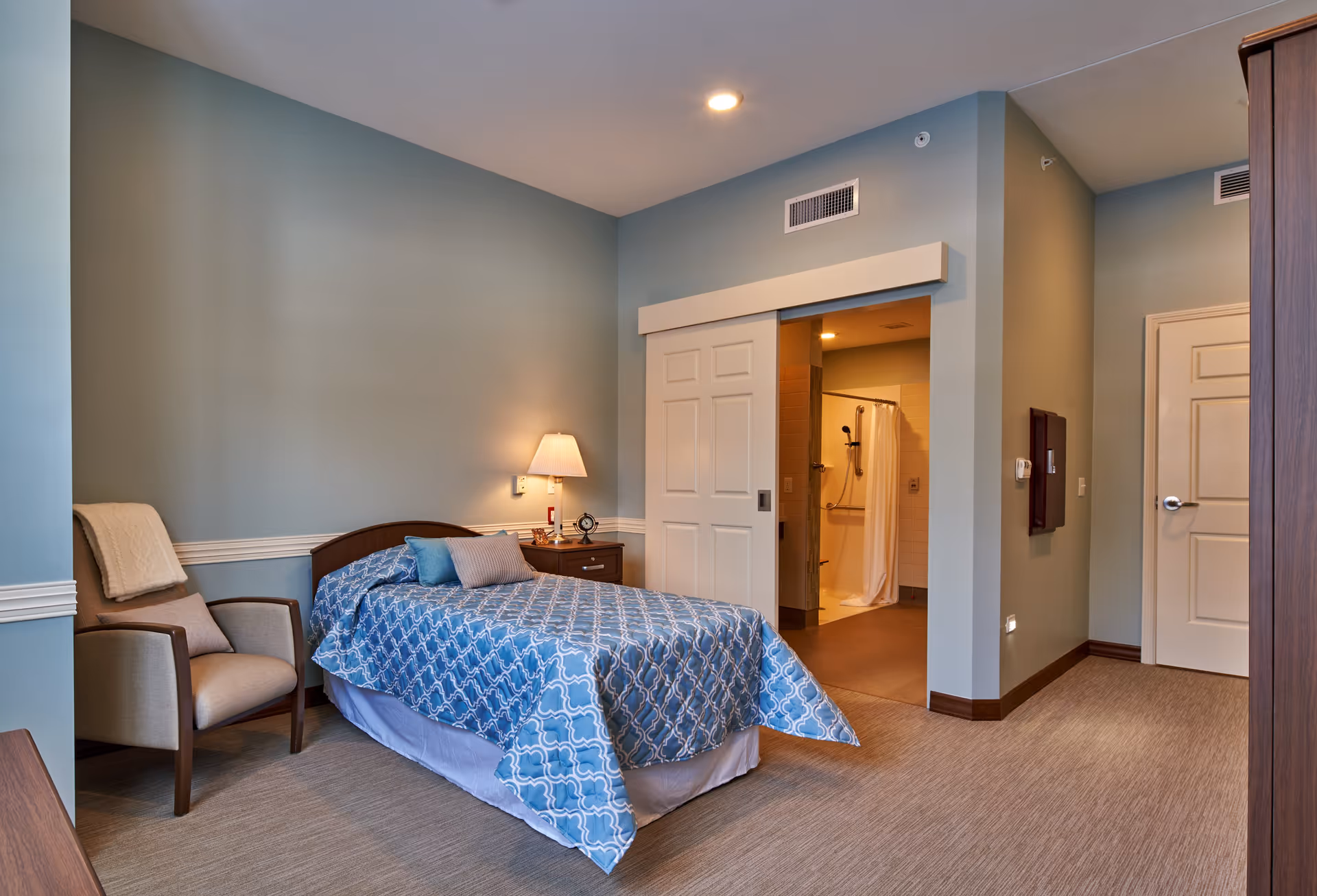 A cozy bedroom with a single bed covered in a blue patterned bedspread, a wooden nightstand with a lamp and clock, and a comfortable armchair with a blanket draped over it. The room has light blue walls and beige carpet. A sliding door is open, revealing a bathroom with a shower and white curtain.