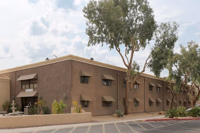 Exterior view of a two-story brown building with multiple windows, some with awnings, surrounded by trees and desert landscaping under a partly cloudy sky.