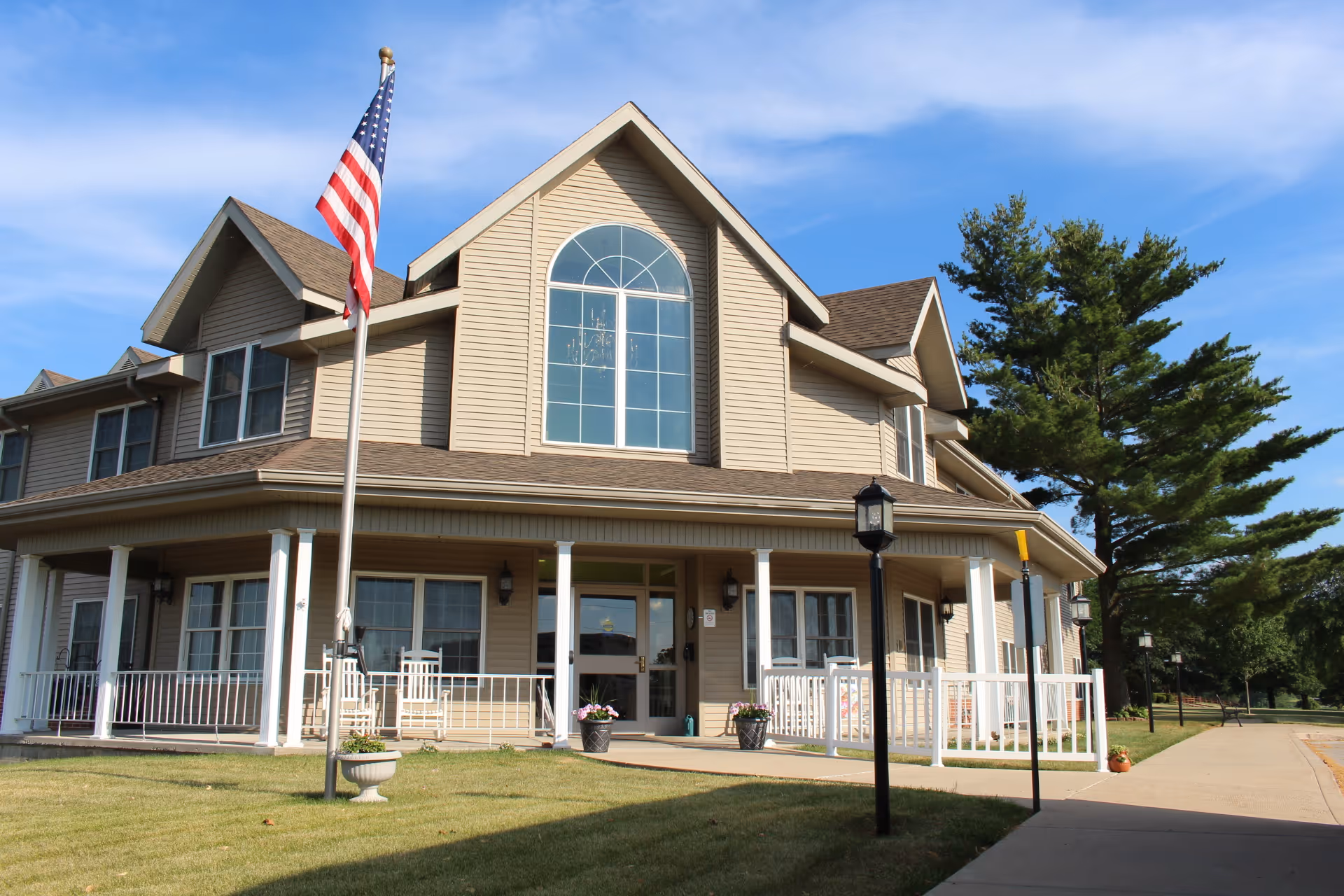 Exterior view of a two-story beige nursing home building with a large arched window above the entrance, a covered porch with white railings and rocking chairs, an American flag on a flagpole in the front yard, and a clear blue sky with some trees in the background.