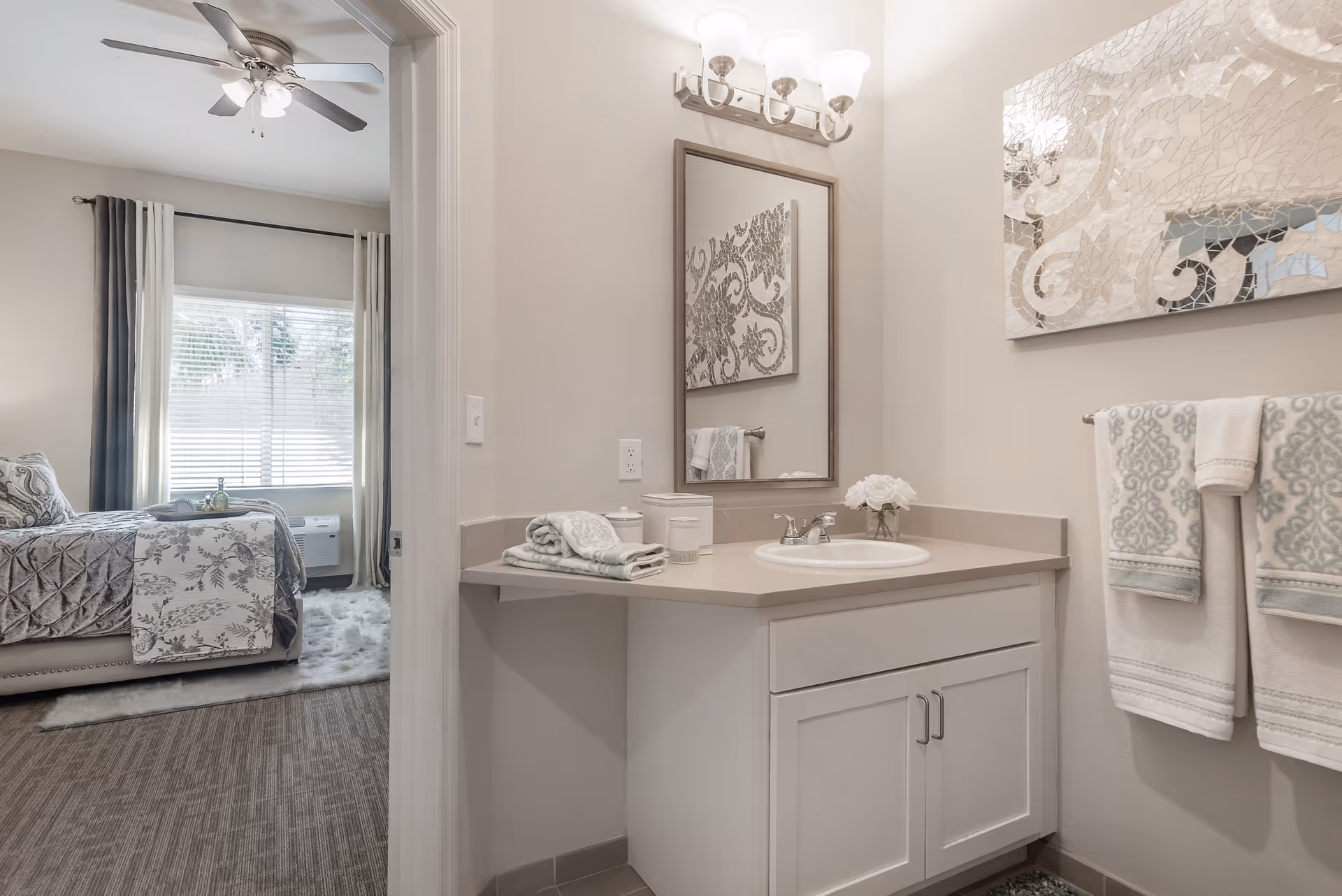 A bathroom vanity area with a sink, mirror, and light fixture above. Towels are neatly folded on the counter and hanging on a towel rack. A decorative piece of art is mounted on the wall. Through an open doorway, a bedroom with a bed, window with curtains, and ceiling fan is visible.