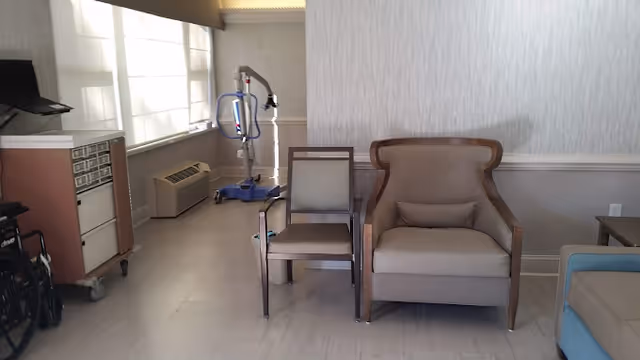 Interior room with two chairs, one wooden with a cushion and one upholstered armchair with a pillow, a wheelchair partially visible on the left, a medical lift device near the window, and a cabinet with drawers. The room has light-colored walls and flooring.