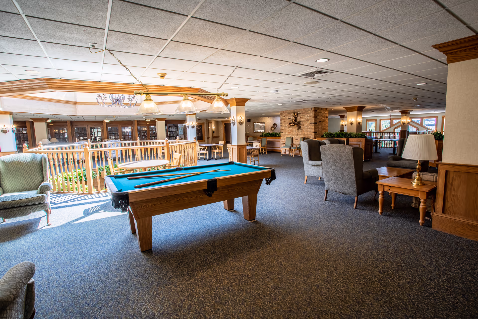 A spacious senior living community lounge area featuring a pool table in the center, surrounded by comfortable armchairs and wooden tables. The room has carpeted floors, a drop ceiling with recessed lighting, and large windows allowing natural light. There is a wooden railing around a sunken area with plants, and a brick fireplace in the background.