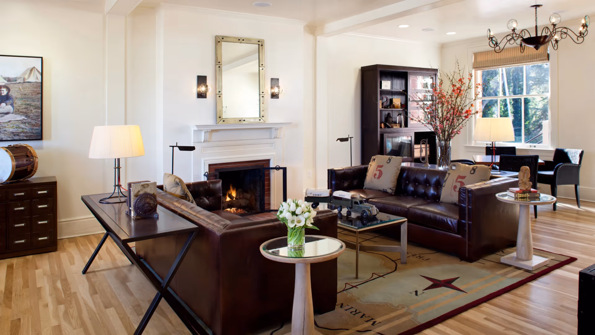 A cozy living room with two dark brown leather sofas arranged around a glass coffee table and a fireplace with a mirror above it. The room features light wooden flooring, a large window with natural light, a chandelier, a dark wooden cabinet, and various decorative items including flowers, books, and a toy truck.