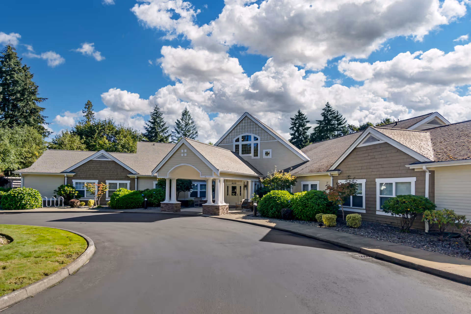 Front entrance of a single-story senior living facility with a covered portico, landscaped shrubs, and a curved driveway under a partly cloudy sky.