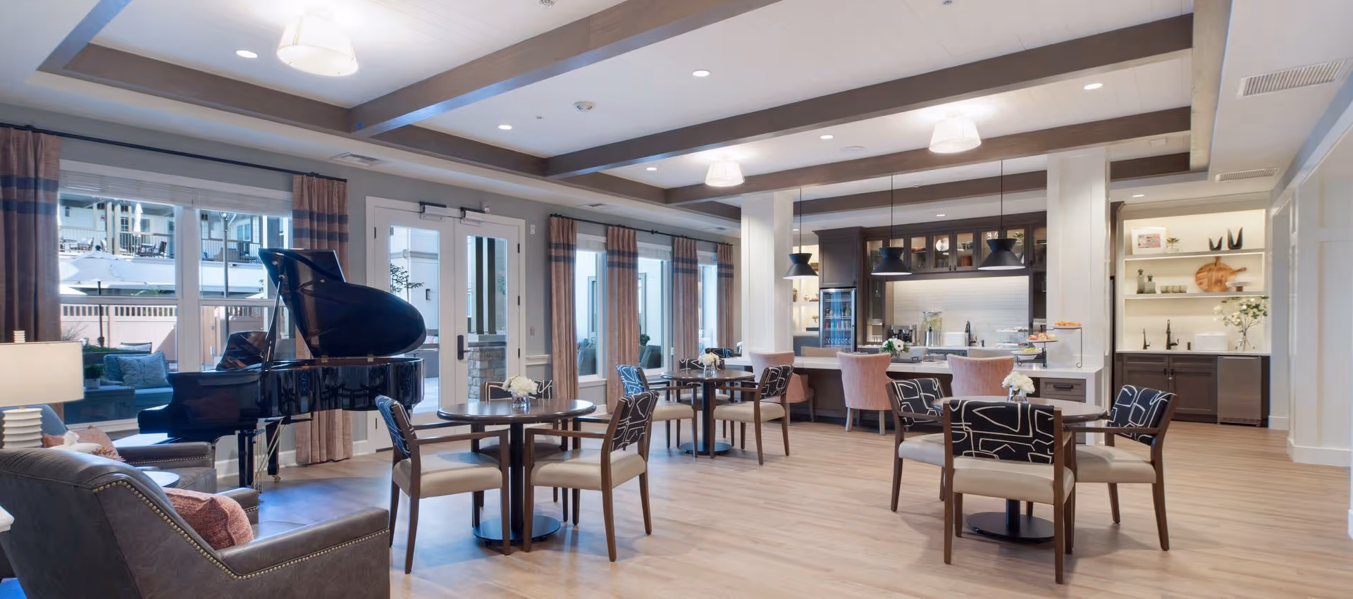 Bright communal dining and lounge area with round tables and chairs, a grand piano, and an open kitchen/bar.