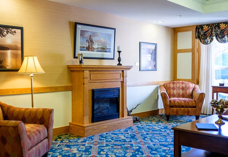 A cozy living room area with two patterned armchairs, a wooden electric fireplace with decorative items on the mantel, framed artwork on the walls, a floor lamp, and a window with floral valance curtains letting in natural light. The room has a blue patterned carpet and light-colored walls with wooden trim.