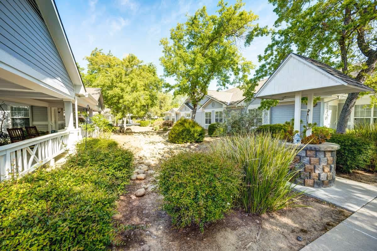 Outdoor view of a senior living facility with light blue buildings, green bushes, trees, and a dry rocky creek bed running through the center. There is a covered well structure on the right and a porch with rocking chairs on the left.