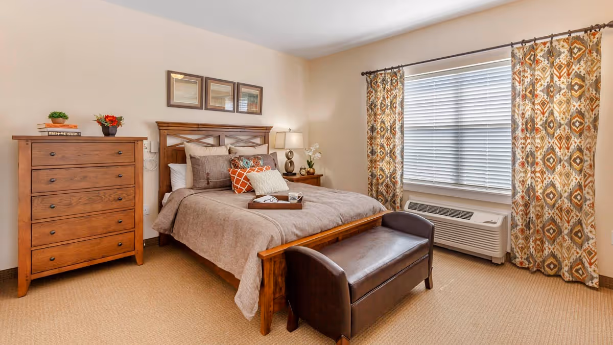 A cozy bedroom with a wooden bed frame and a neatly made bed featuring multiple pillows and a tray with a cup and napkins. There is a wooden chest of drawers with decorative plants and books on top, a bedside table with a lamp and a small plant, patterned curtains framing a window with blinds, and a cushioned bench at the foot of the bed.