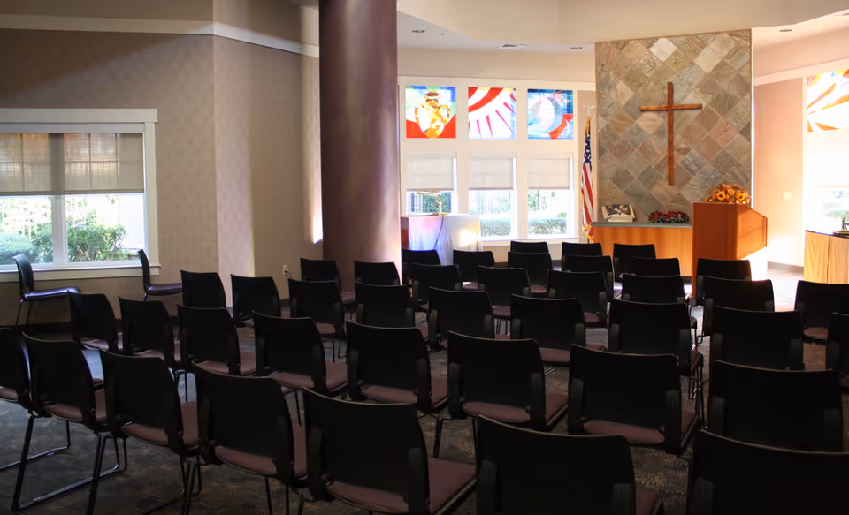 Interior view of a chapel or meeting room with rows of black chairs facing a podium and a wall with a large wooden cross mounted on stone tiles. Stained glass windows and an American flag are visible in the background.
